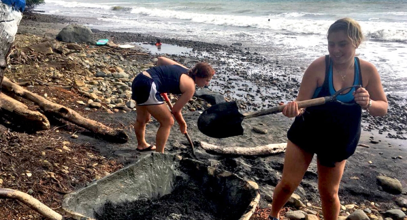 two people work on a beach, using shovels and a wheelbarrow, during a service project